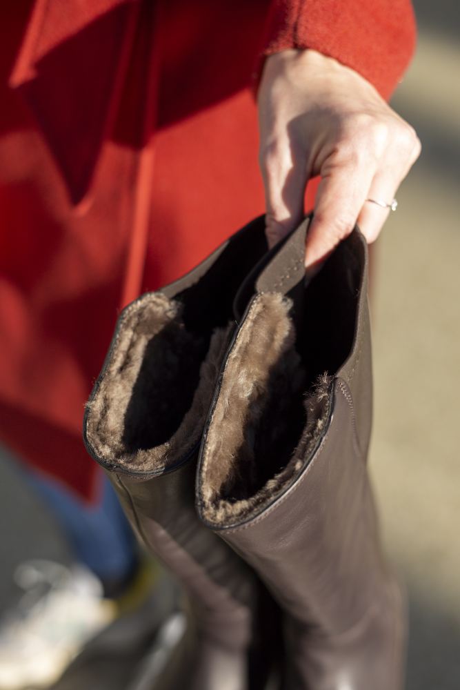 Eine Person in einem roten Mantel hält ein Paar ZAQQ QATCH Winter Brown-Stiefel aus Nappaleder mit Lammfell-Futter und präsentiert ihr kuscheliges Innenleben in Nahaufnahme. Die Stiefel werden im Freien gehalten, im Hintergrund ist ein Sneaker zu sehen.