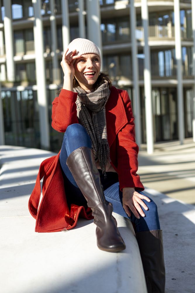 Eine Person mit einem roten Mantel, einer weißen Beanie und den QATCH Nappa Brown-Stiefeln von ZAQQ sitzt an einem sonnigen Tag im Freien auf einer Betonfläche, lächelt und blickt in die Ferne. Im Hintergrund sind hohe Gebäudestrukturen zu sehen.