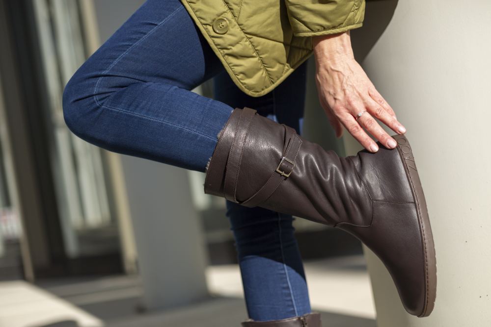 Eine Person trägt die braunen Lederstiefel ATTRAQT Winter von ZAQQ, kombiniert mit Blue Jeans und einem grünen Mantel. Sie lehnt an einer Stange und richtet ihre Stiefel. Die Szene spielt im Freien vor der Kulisse eines modernen Gebäudes.