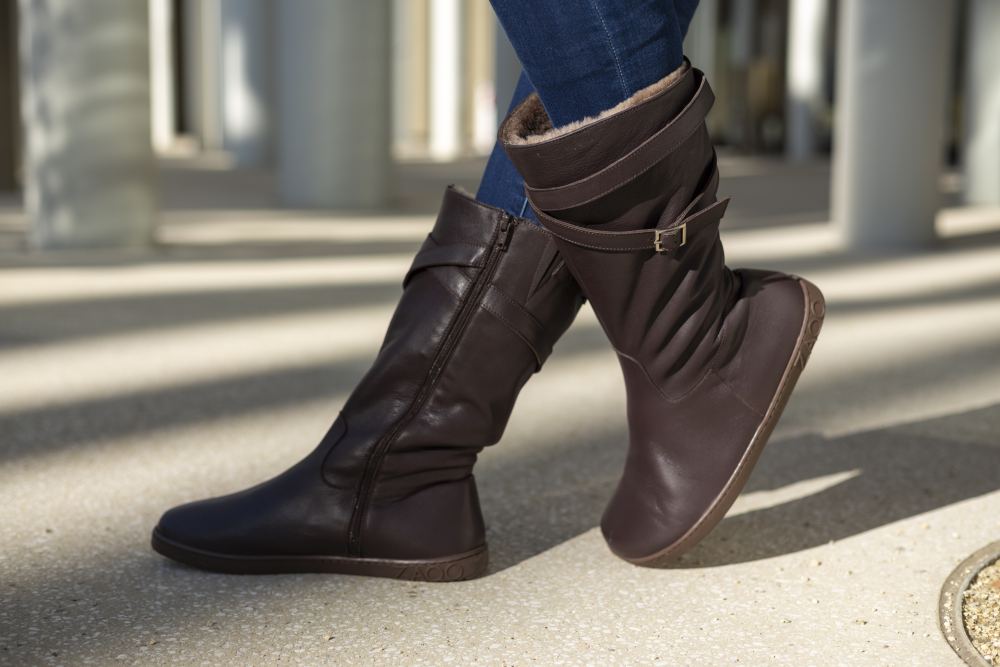 Eine Person trägt die stylischen dunkelbraunen ATTRAQT Winter Brown Kniestiefel von ZAQQ mit Schnallendetail. Diese Stiefel sind für ihren außergewöhnlichen Tragekomfort bekannt, aus einem glatten Material gefertigt und mit einem seitlichen Reißverschluss ausgestattet. Sie werden mit dunkelblauen Jeans vor einem sanft verschwommenen Hintergrund kombiniert, der helle Säulen und Schatten zeigt.