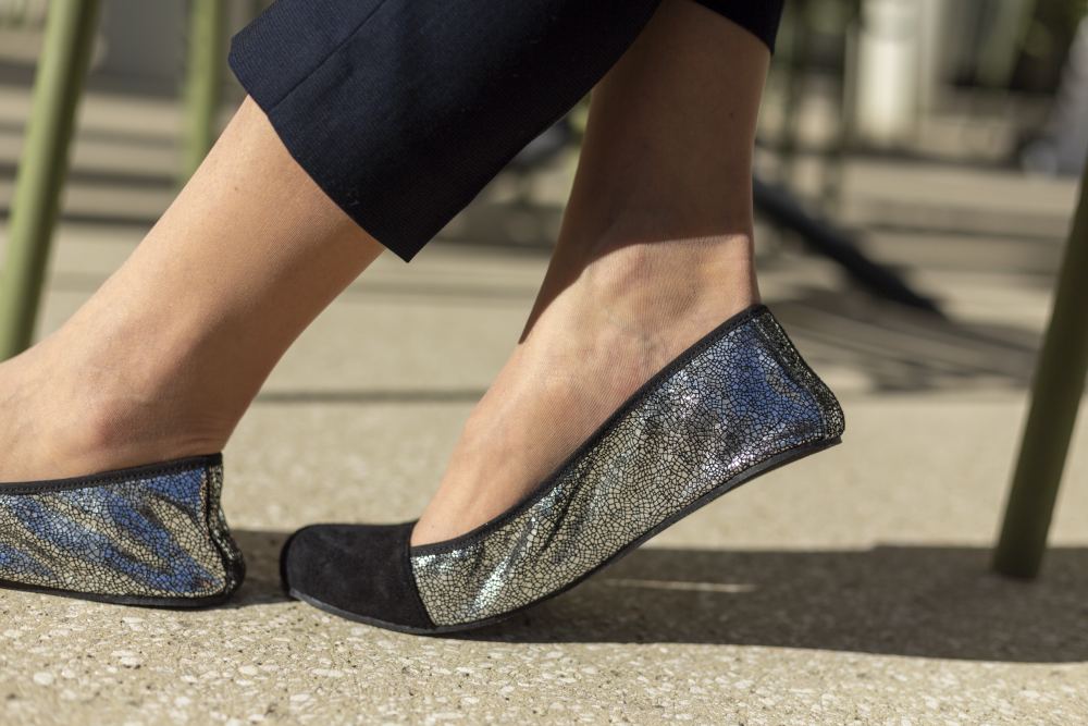 Eine Person in den Schuhen TWIST Derby Black von ZAQQ, die metallisch glitzernde Oberflächen mit schwarzen Zehenkappen aufweisen und im Stil von Ballerinaschuhen gehalten sind, steht in schwarzer Hose auf einer sonnenbeschienenen Steinfläche. Im Hintergrund sind grüne Stuhlbeine aus Metall zu sehen.