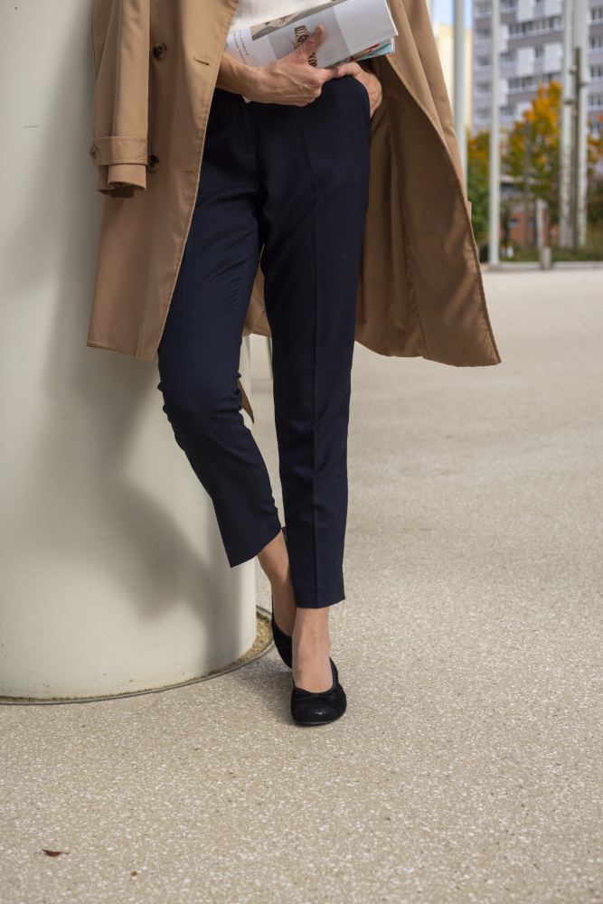 Eine Person in einem beigen Trenchcoat und marineblauen Hosen steht an einer weißen Säule und hält eine Zeitschrift in der Hand. Ihr Gesicht ist nicht zu sehen und sie trägt schwarze TWIST Velours-Schuhe von ZAQQ. Der urbane Hintergrund mit Gebäuden und Bäumen in der Ferne unterstreicht die lebendige Flexibilität der Stadt.