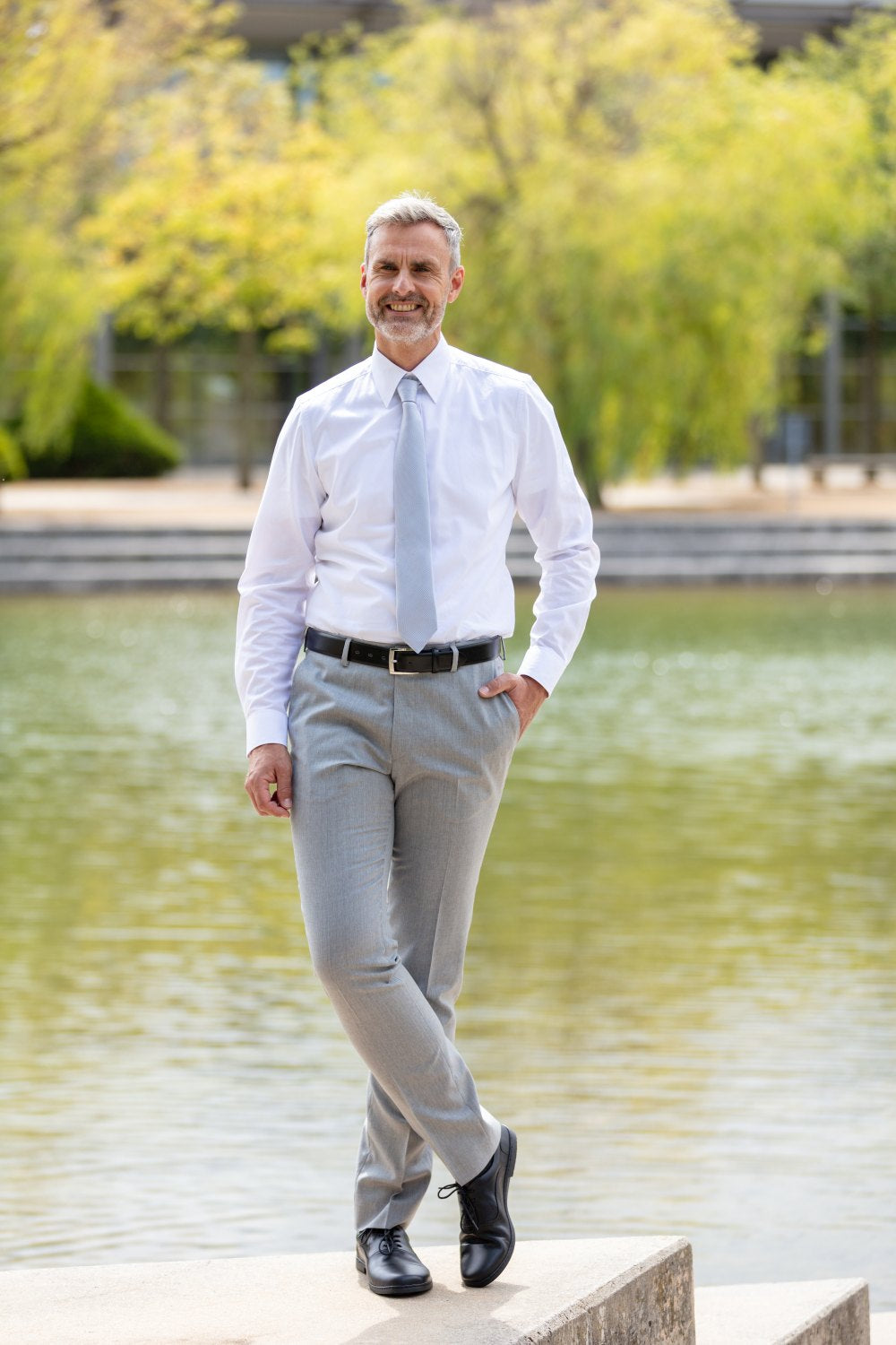 Ein Mann mit grauem Haar und Schnurrbart steht neben einem Gewässer. Er trägt ein weißes Hemd, eine hellgraue Krawatte und graue Hosen. Er stützt sich auf ein Bein, trägt stylische schwarze ZAQQ APEQ-Schuhe und lächelt in einer Parklandschaft mit Grün im Hintergrund.