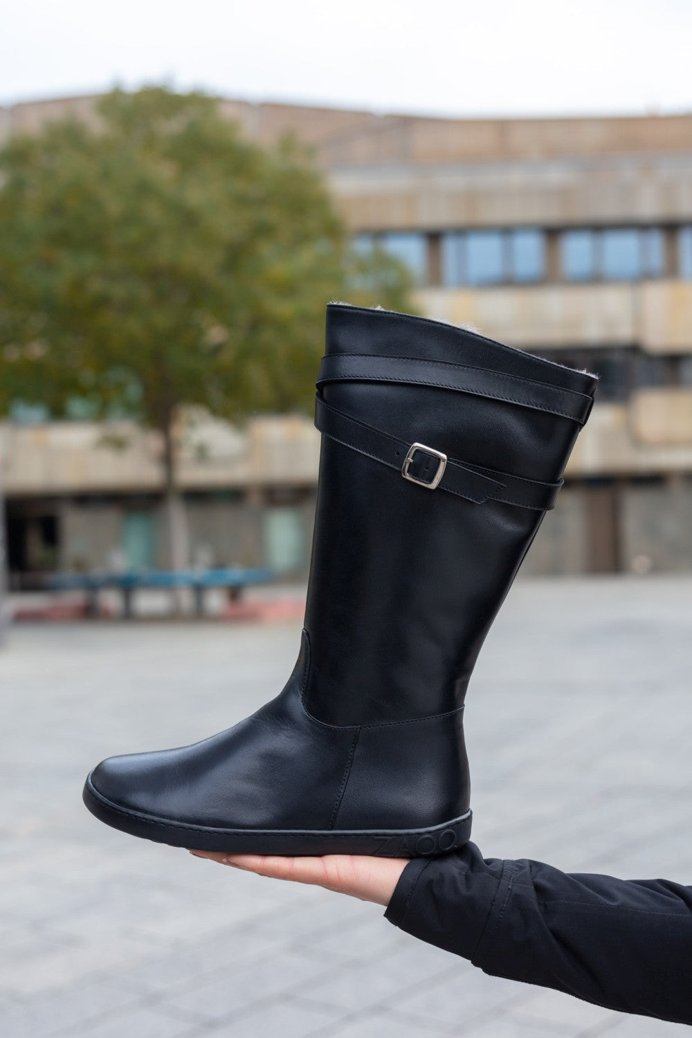 In einer Outdoor-Umgebung hält eine Person den eleganten ATTRAQT Winter Black-Stiefel von ZAQQ, gefertigt aus Ziegennappaleder. Der Stiefel hat einen dezenten Schnallenriemen in der Nähe der Oberseite, mit Bäumen und einem sanft verschwommenen Gebäude im Hintergrund.