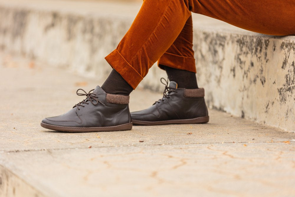 Eine Person sitzt in der Hose NEWQUAY Brown und den stylischen Schuhen ZAQQ auf einer Steintreppe, wobei der Fokus auf den Beinen und dem eleganten Schuhwerk liegt.