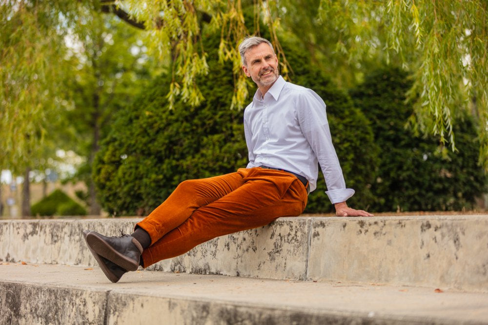 Ein Mann mit ergrauendem Haar sitzt auf einer niedrigen Steinmauer in einem Park. Er trägt ein weißes Hemd und orangefarbene Hosen. Er trägt die Schuhe NEWQUAY Brown von ZAQQ, die aus geschmeidigem Nappaleder gefertigt sind. Im Hintergrund sind üppige grüne Bäume und Blätter zu sehen, die eine ruhige Außenkulisse schaffen.