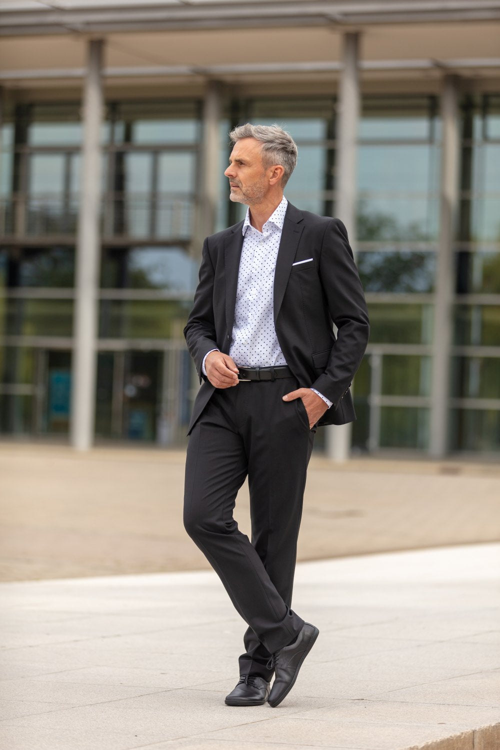 Ein Mann mit grauem Haar, gekleidet in einen schwarzen Anzug und ein gemustertes Hemd, steht im Freien und trägt PEAQ Black-Schuhe von ZAQQ mit den Händen in den Taschen. Als Hintergrund dient ein modernes Glasgebäude.