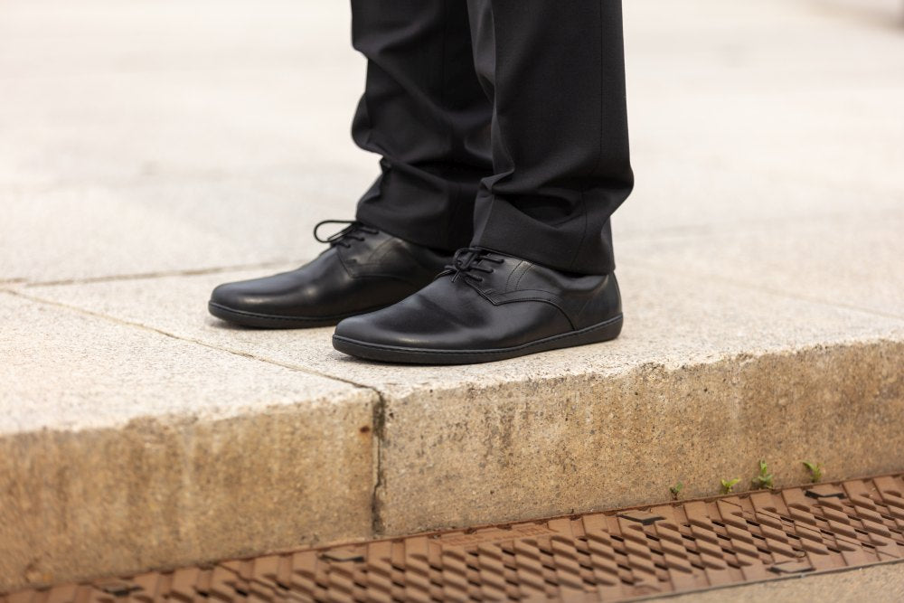 Eine Person steht in schwarzen Abendschuhen und einer schwarzen Hose auf einer Betonstufe. Die eleganten PEAQ Black-Schuhe von ZAQQ mit bequemen Sohlen sind ordentlich geschnürt und stehen auf einem Gehweg oder Bürgersteig.