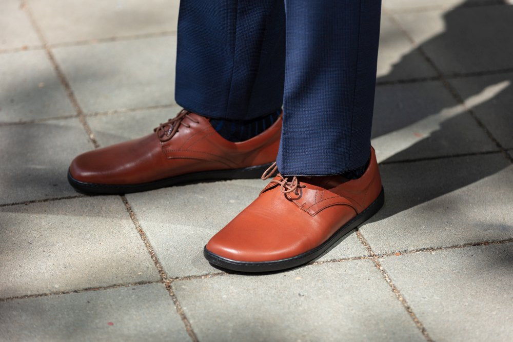 Eine Person in ZAQQs PEAQ Cognac Lederschuhen und marineblauer Hose steht auf grauen Gehwegplatten. Das glatte Glattleder glänzt dezent im Licht und wirft elegante Schatten auf den Boden.