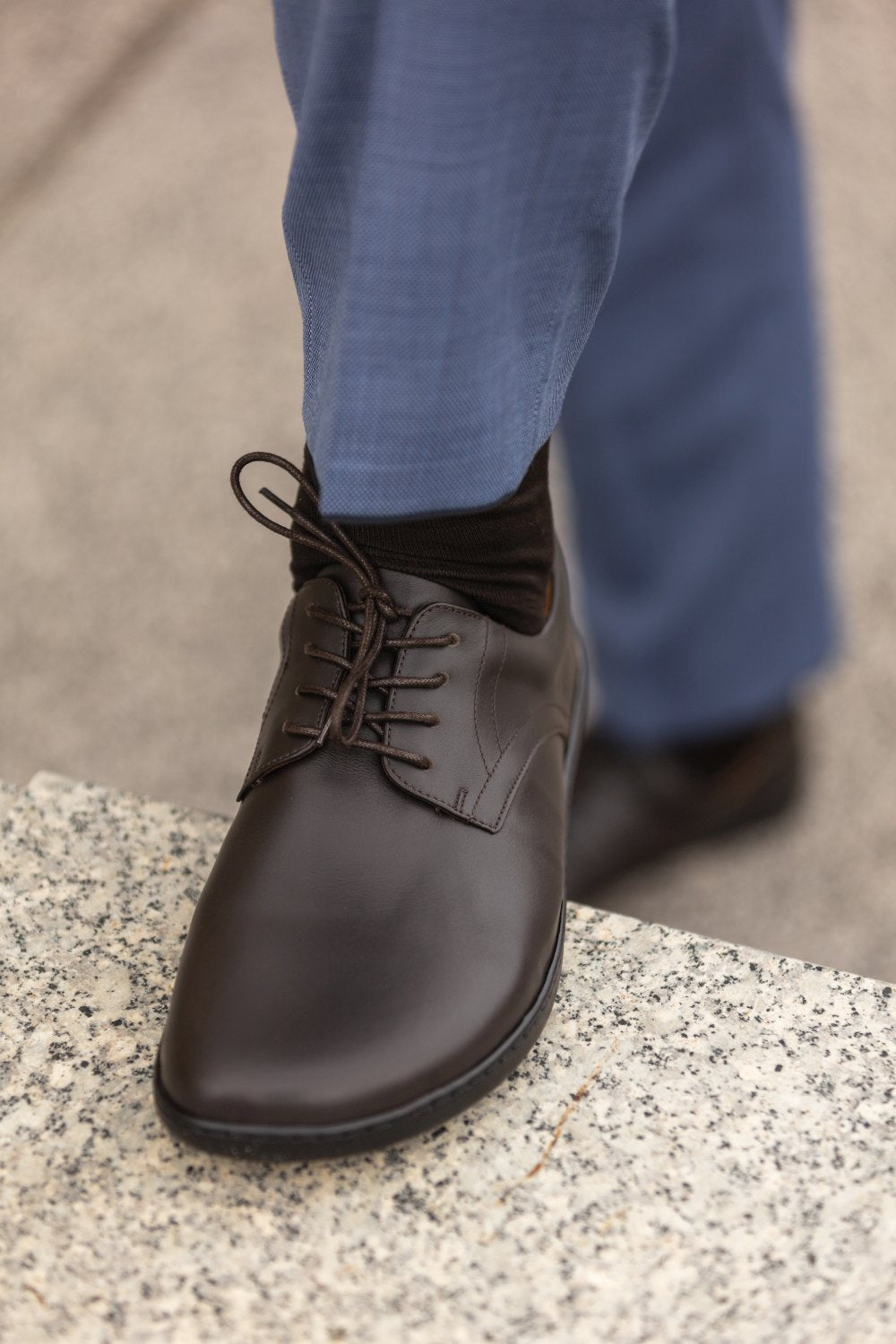 Eine Nahaufnahme einer Person, die den PEAQ Dark Brown von ZAQQ mit schwarzen Schnürsenkeln trägt und auf eine Granitoberfläche tritt. Sie trägt blaue Hosen und schwarze Socken und zeigt damit einen Business-Casual-Stil.