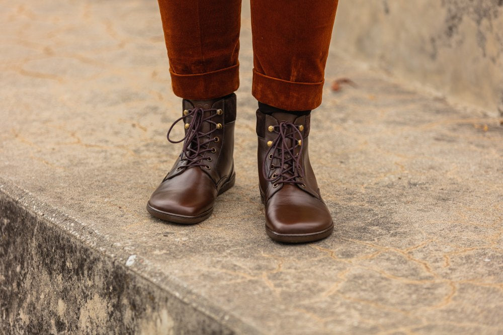 Eine Person steht auf einer strukturierten Steinoberfläche und trägt die QORBY Brown-Stiefel von ZAQQ, deren anschmiegsame Passform die rostfarbene Hose perfekt ergänzt. Die Hose ist an den Bündchen leicht hochgekrempelt, was die volle Höhe dieser halbhohen Stiefel betont.