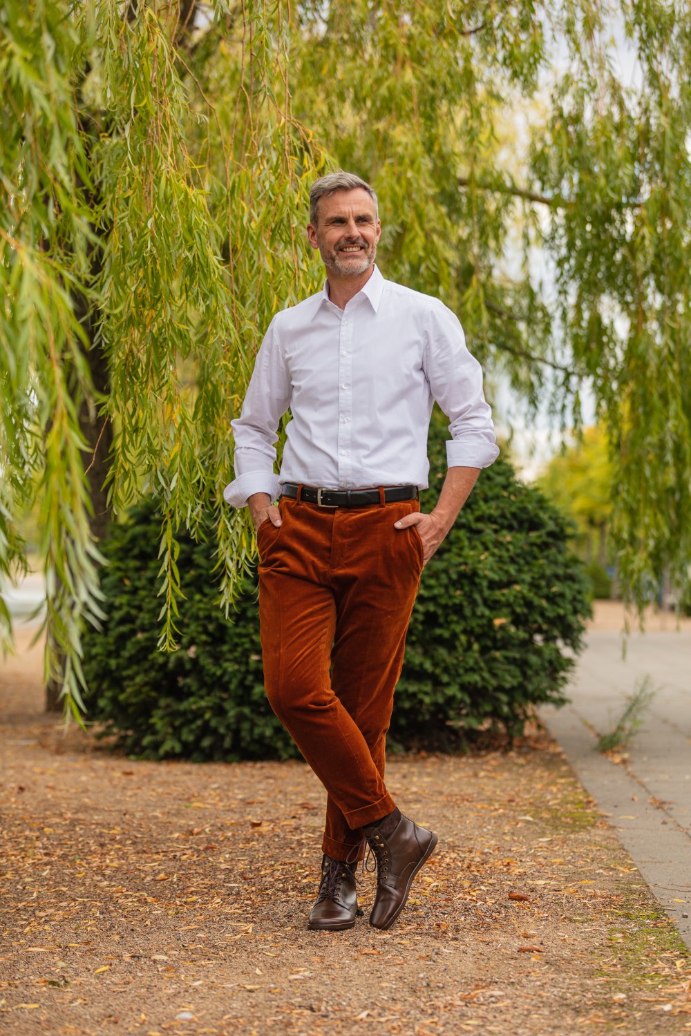 Ein Mann in weißem Hemd und rostfarbener Hose steht lässig mit den Händen in den Taschen unter einem Baum mit herabhängenden Ästen. Er lächelt und trägt braune QORBY-Stiefel von ZAQQ. Im Hintergrund sind ein gepflasterter Weg und Grünflächen zu sehen.