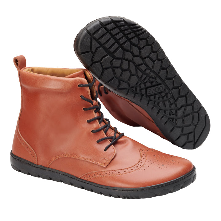 Ein Paar QUINTIC Brogue Cognac von ZAQQ mit braunem Leder und schwarzen Sohlen. Ein Stiefel steht aufrecht und zeigt aufwendige Zehennähte, während der andere auf der Seite liegt und das strukturierte Profil und eine Aufreißlasche an der Rückseite hervorhebt.