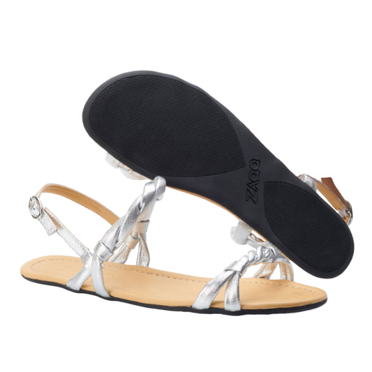 Ein Paar ZAQQ QLITTER Silver-Sandalen, von denen eine flach liegt und die andere die schwarze Sohle betont, verfügen über verstellbare Fesselriemen und ein minimalistisches Design, komplett mit einem hellbraunen Fußbett – ideal für einen eleganten Abend.