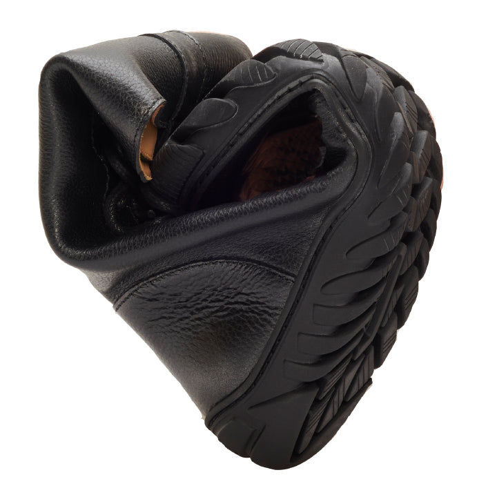 Ein schwarzer Schuh mit strukturierter ULTRAGRIP-Sohle ist gebogen, um seine Flexibilität zu demonstrieren. Der schwarze Schuh ZAQQ DOQS ist teilweise gefaltet, wodurch sein geschmeidiges Premium-Nappaleder-Material und sein Design hervorgehoben werden.