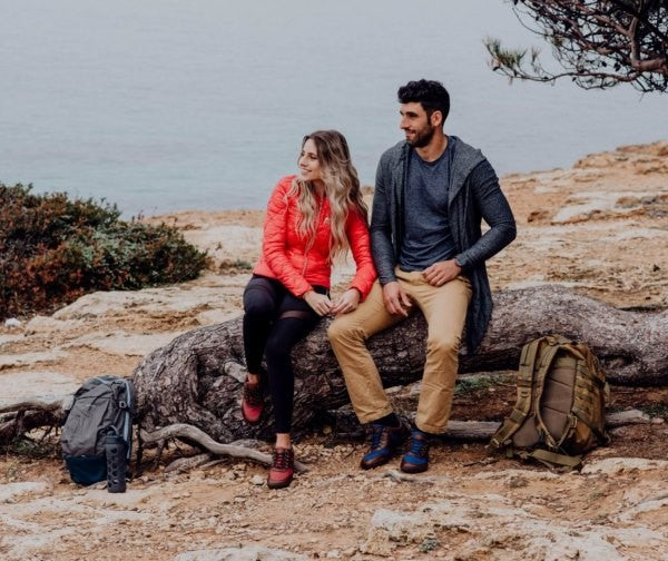 Eine Frau in einer roten Jacke und ein Mann in einer grauen Strickjacke sitzen auf einem großen Stück Treibholz an einer felsigen Küste. Sie sind von spärlicher Vegetation umgeben. In der Nähe stehen zwei Rucksäcke. Im Hintergrund ist das Meer unter einem bewölkten Himmel zu sehen.