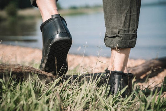 Nahaufnahme von zwei Personen, die auf Gras in der Nähe eines Gewässers laufen. Eine Person trägt schwarze knöchelhohe Stiefel und hochgekrempelte dunkelgrüne Hosen. Der Blick konzentriert sich auf ihre Unterschenkel und Schuhe, im Hintergrund sind ein Holzstamm und Kieselsteine zu sehen.