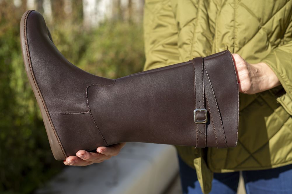 Eine Person steht im Freien und hält den ZAQQ ATTRAQT Vegan Brown mit Schnallendetail, das die flexible Sohle hervorhebt. Sie trägt eine grüne Steppjacke, perfekt für das Wetter.