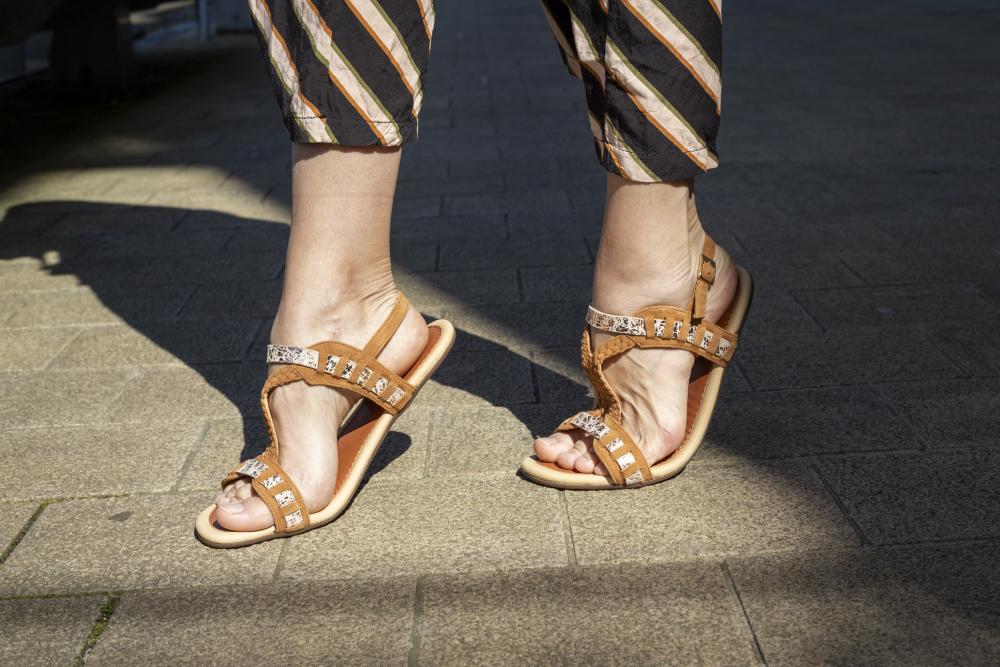 Nahaufnahme einer Person, die die BLINQ Brown Sandalen von ZAQQ mit eleganten silberfarbenen Riemchen trägt, während sie auf einem sonnenbeschienenen Ziegelsteinpflaster steht. Das feminine Design ergänzt perfekt ihre gestreifte Hose für einen mühelos schicken Look.