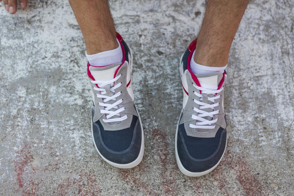 Nahaufnahme von zwei Füßen in dunkelblauen BREAQ-Sneakern von ZAQQ mit weißen Schnürsenkeln, die auf einer strukturierten Betonoberfläche stehen. Die Person trägt weiße Söckchen, die dem Sneaker-Look einen frischen Akzent verleihen.