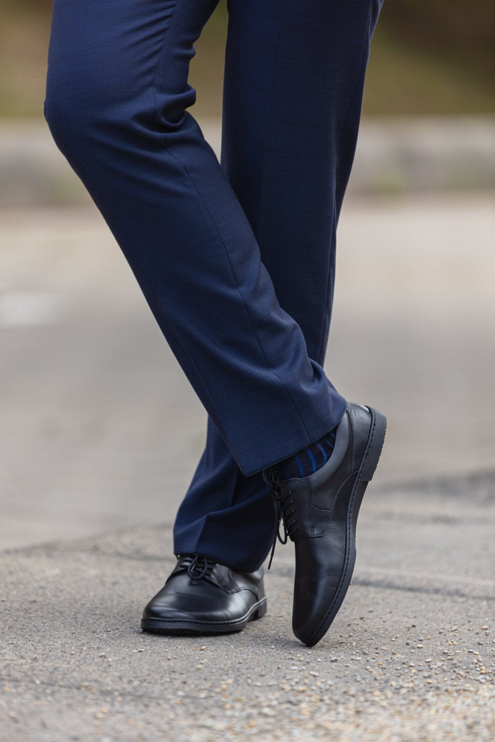 Eine Person in marineblauen Hosen und polierten BRIQ Black-Schuhen von ZAQQ steht mit gekreuzten Beinen auf einer gepflasterten Fläche. Diese schwarzen Abendschuhe mit zeitlosem Design passen gut zu dezent gemusterten Socken.