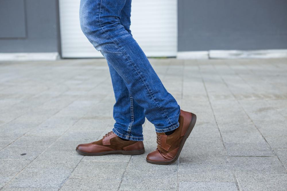 Eine Person in Blue Jeans und BRIQ Brogue Antique Cognac-Schuhen von ZAQQ läuft auf einem grau gepflasterten Gehweg. Im Hintergrund sind eine graue Wand und ein weißer Fensterladen zu sehen.