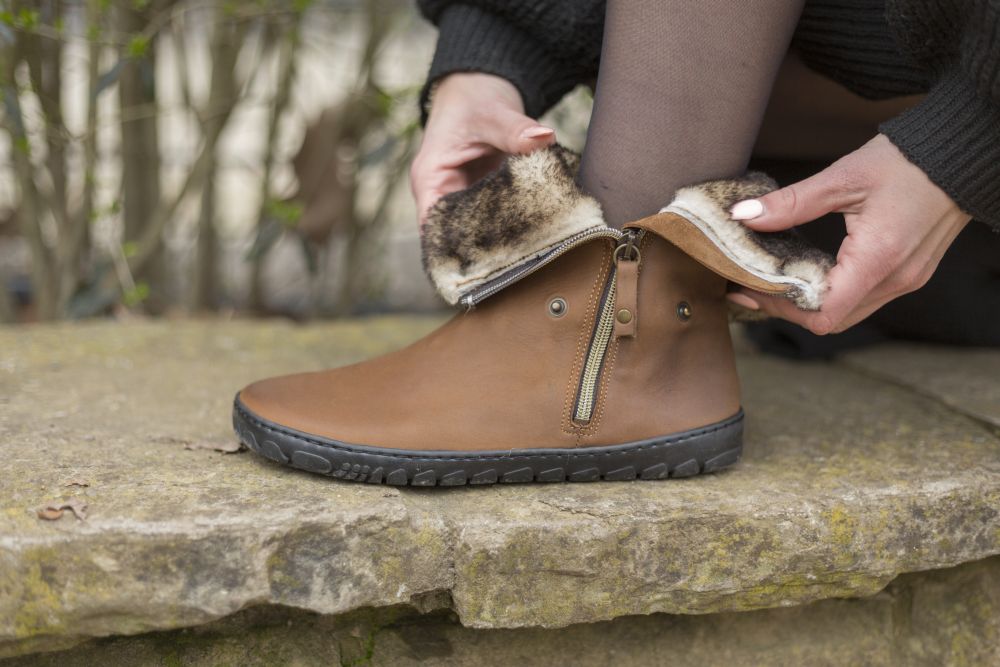 Eine Person sitzt auf einer Steinoberfläche und passt einen ZAQQ HUQ braunen Stiefel mit sichtbarem Reißverschluss und pelzigem Futter an. Der Stiefel hat eine strukturierte schwarze Kautschuksohle und die Person trägt dunkle Strumpfhosen und einen schwarzen Ärmel. Ein verschwommener Hintergrund aus Veloursleder-Gebüsch vervollständigt die Szene.