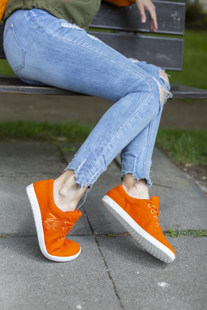 Eine Person sitzt auf einer Bank im Freien. Sie trägt eine Jeans im Used-Look und die leuchtenden LIQE Orange Sneaker von ZAQQ, die für ein Barfußgefühl sorgen. Der gepflasterte Boden kontrastiert mit dem grünen Gras im Hintergrund.