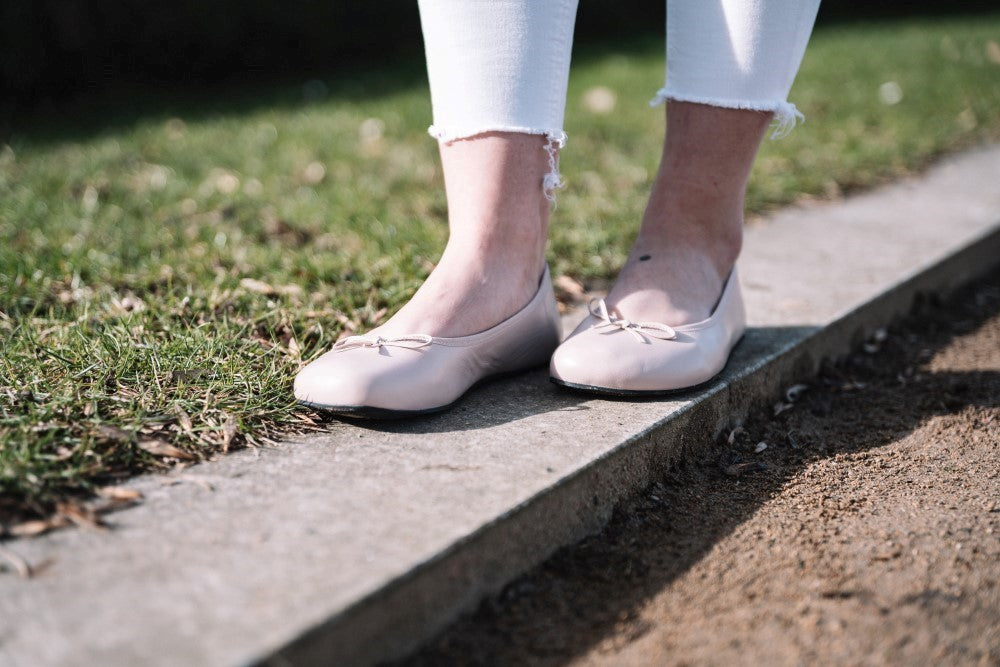 Eine Person steht auf einem schmalen Betonweg, auf dessen einer Seite Gras und auf der anderen Erde liegt. Sie trägt elegante LOOQ Nude Ballerinas von ZAQQ in Hellrosa. Die Person trägt weiße Hosen mit ausgefransten Säumen.