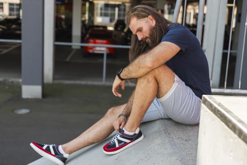 Ein Mann mit langen Haaren sitzt auf einem städtischen Felsvorsprung und bindet die Schnürsenkel seiner roten OAQLEY-Sneaker von ZAQQ. Er trägt ein marineblaues Hemd und hellgraue Shorts und trägt eine Uhr an seinem linken Handgelenk in einer legeren Outdoor-Situation.