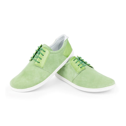 Ein Paar PIQUANT Lime Casual-Sneaker von ZAQQ aus Nappa- und Wildleder mit weißer Sohle und grünen Schnürsenkeln. Ein Barfuß-Sneaker lehnt an dem anderen vor einem makellos weißen Hintergrund.