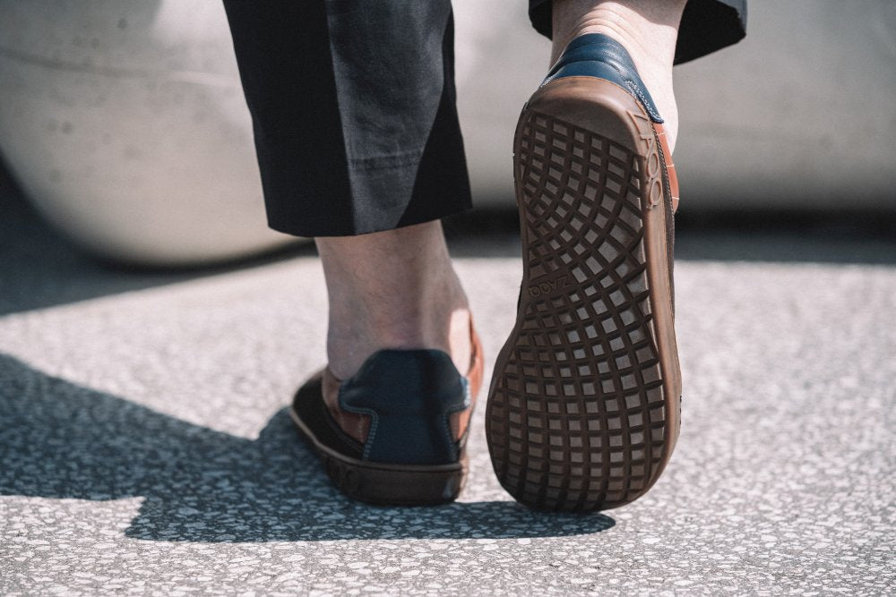 Das Bild zeigt eine Nahaufnahme einer Person, die auf einer strukturierten Oberfläche läuft. Sie trägt schwarze Hosen und die stylischen QAMPION Brown Loafer von ZAQQ. Der Fokus liegt auf der Sohle eines Schuhs, die ihr kompliziertes Muster aus flexibler Kautschuksohle zeigt, das für Komfort bei jedem Schritt sorgt.