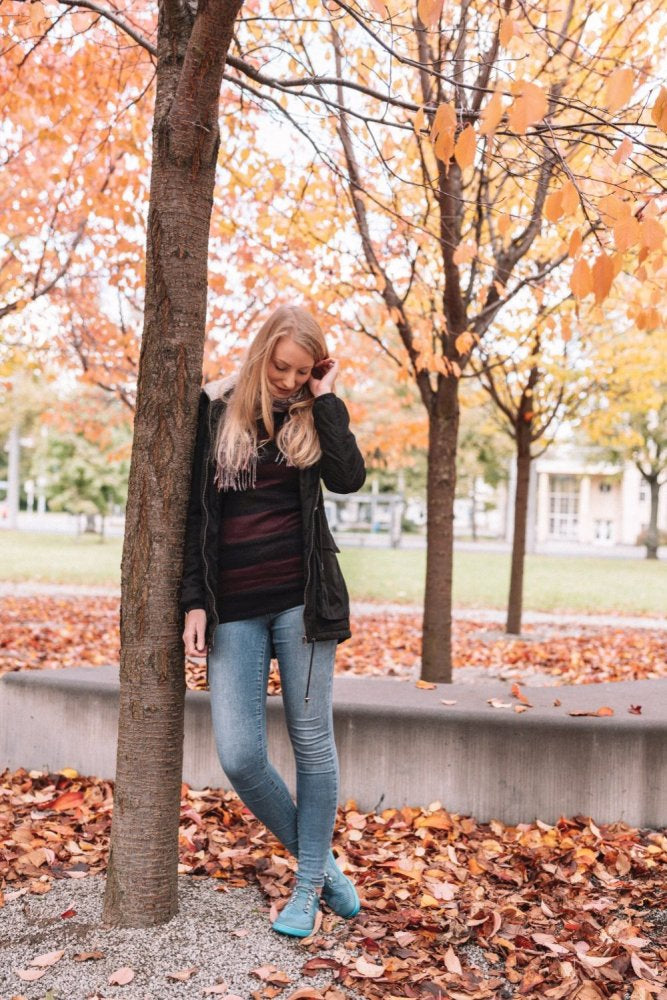 Eine Person mit langen Haaren und einer schwarzen Jacke lehnt an einem Baum in einem Park, ihre QARLY Turquoise Sneaker von ZAQQ streifen das Herbstlaub. Die Bäume sind mit leuchtend orangefarbenem Laub bedeckt, was einen markanten Kontrast bildet, der die besinnliche Aura der Szene verstärkt.