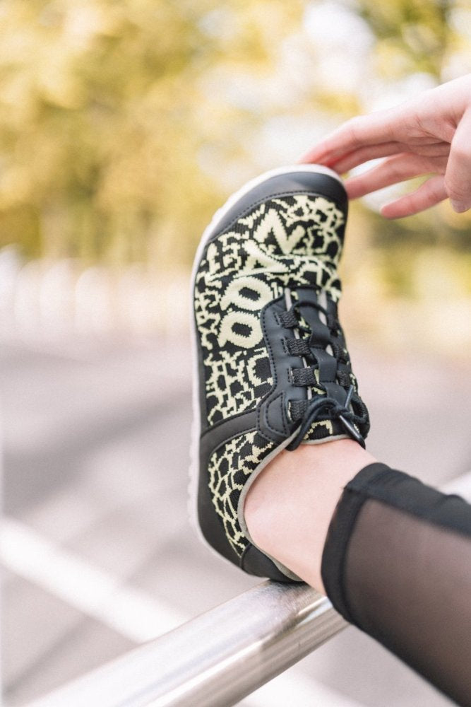 Eine Person, die die QASEY Green-Sneaker von ZAQQ mit einem schwarz-grünen abstrakten Muster trägt, stellt einen Fuß auf ein Metallgeländer. Diese Schuhe sind aus veganen Materialien und atmungsaktivem Netzgewebe gefertigt und für mehr Komfort geschnürt. Im Hintergrund ist eine verschwommene Außenkulisse mit viel Grün zu sehen.