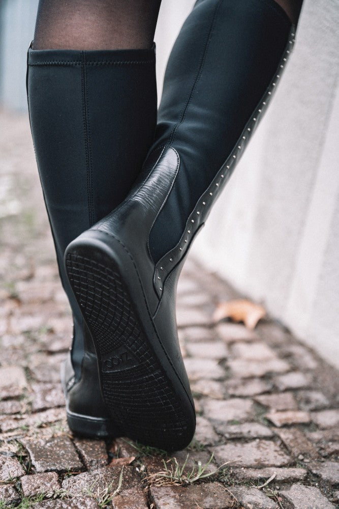 Nahaufnahme einer Person, die die hohen schwarzen QATCH SHINE Nappa-Stiefel von ZAQQ auf einem Kopfsteinpflasterweg trägt. Die Stiefel haben ein glänzendes Seitenteil und Nietendetails entlang der hinteren Naht. In der Nähe liegt ein einzelnes gefallenes Blatt auf dem Boden.