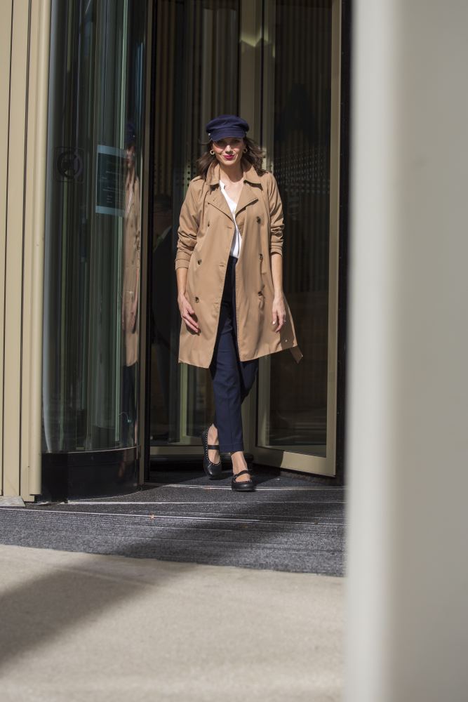 Eine Person in einem hellbraunen Trenchcoat, marineblauen Hosen und eleganten schwarzen DIEQO Nappa Boots von ZAQQ, die die natürliche Bewegung der Füße fördern, verlässt ein Gebäude mit Glastüren. Eine marineblaue Kappe und eine Sonnenbrille vervollständigen den Look, als sie an einer weißen Säule im Hintergrund vorbeigehen, die die Sicht teilweise verdeckt.