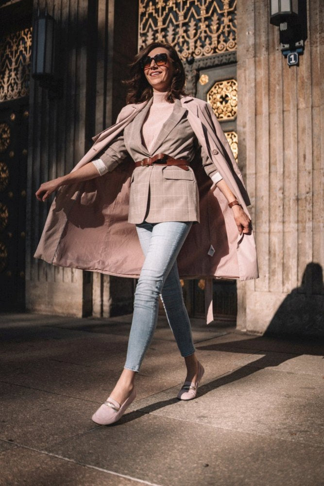 Eine stilvolle Frau schreitet selbstbewusst eine sonnenbeschienene Straße entlang. Ihr Veloursledergürtel hebt ihren karierten Blazer hervor. Mit einer Sonnenbrille schützt sie ihre Augen. Zu hellblauen Jeans trägt sie passende rosa QERP Candy-Schuhe von ZAQQ mit einer strapazierfähigen Kautschuksohle, alles unter einem schicken Trenchcoat. Verzierte Tore und Steinsäulen bilden den Hintergrund.