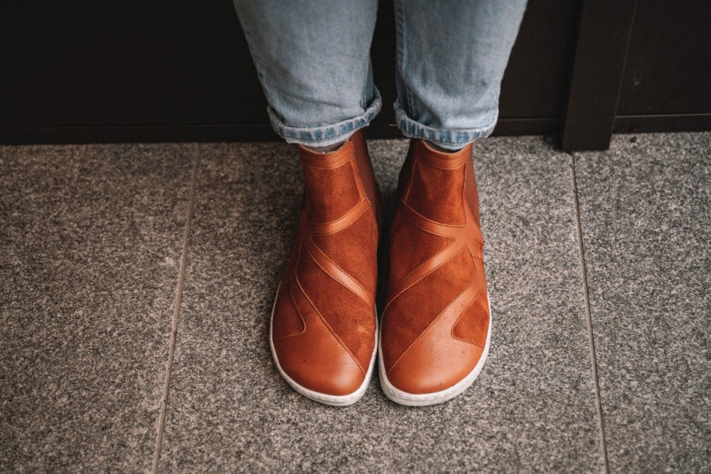 Eine Person, die ZAQQs QEY WEST Cognac-Stiefel und hellblaue Jeans trägt, steht auf einem gesprenkelten grauen Boden. Diese stylischen Stiefel zeichnen sich durch ein modernes Design mit kreuz und quer verlaufenden Mustern aus und sind somit perfekt für jeden lässigen Ausflug.