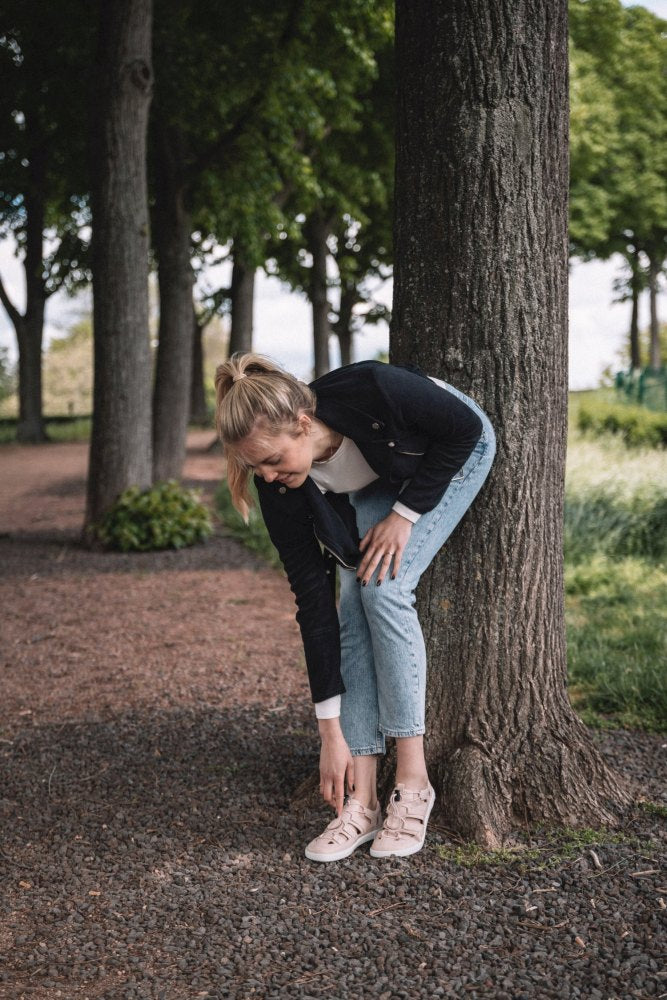 Eine Frau mit blonden Haaren, bekleidet mit einer schwarzen Jacke, einem weißen Hemd und Jeans, bückt sich, um ihr QLEAR Light Pink von ZAQQ zu richten, während sie auf einem von Bäumen gesäumten Kiesweg an einem Baum lehnt.