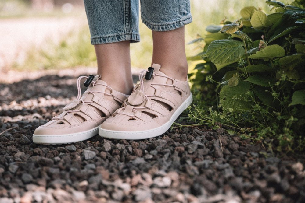 Eine Person, die die QLEAR Light Pink-Sandalen von ZAQQ trägt, steht auf einem Kiesweg. Die Sandalen haben ein Gitterdesign mit weißen Sohlen für besseren Halt. Ihre Jeans sind leicht gekürzt und auf der rechten Seite sind grüne Blätter zu sehen.