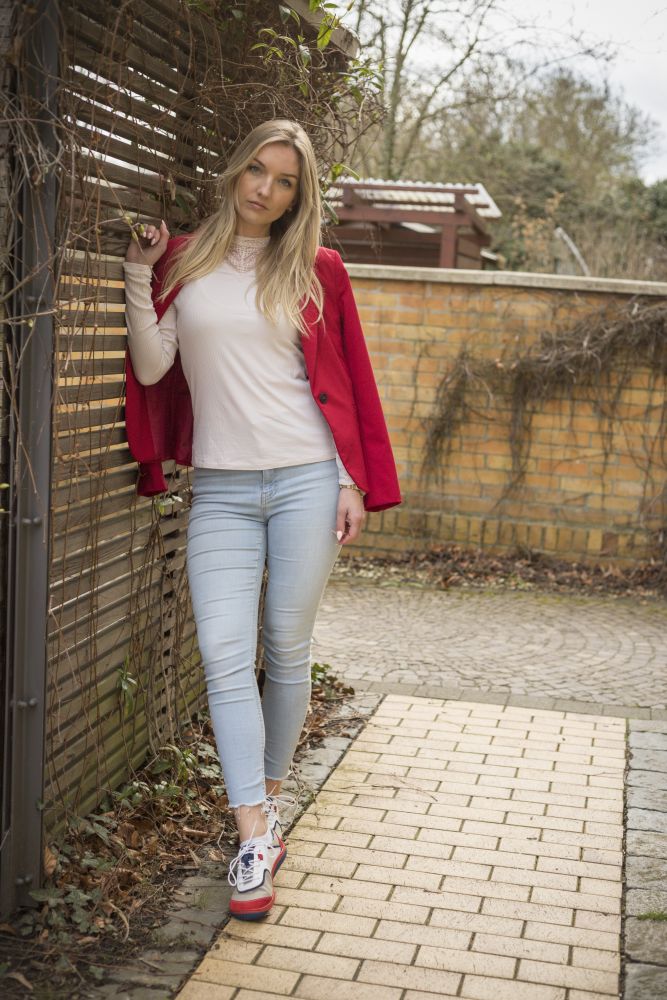 Eine Frau mit langen blonden Haaren, bekleidet mit einer roten Jacke über einem weißen Oberteil und hellblauen Jeans, steht im Freien. Sie trägt den stylischen ZAQQ Damen Sneaker QOLETTE White und lehnt an einem Holzzaun auf einem mit Ziegeln gesäumten Weg. Im Hintergrund sind eine Ziegelmauer und einige Äste zu sehen.