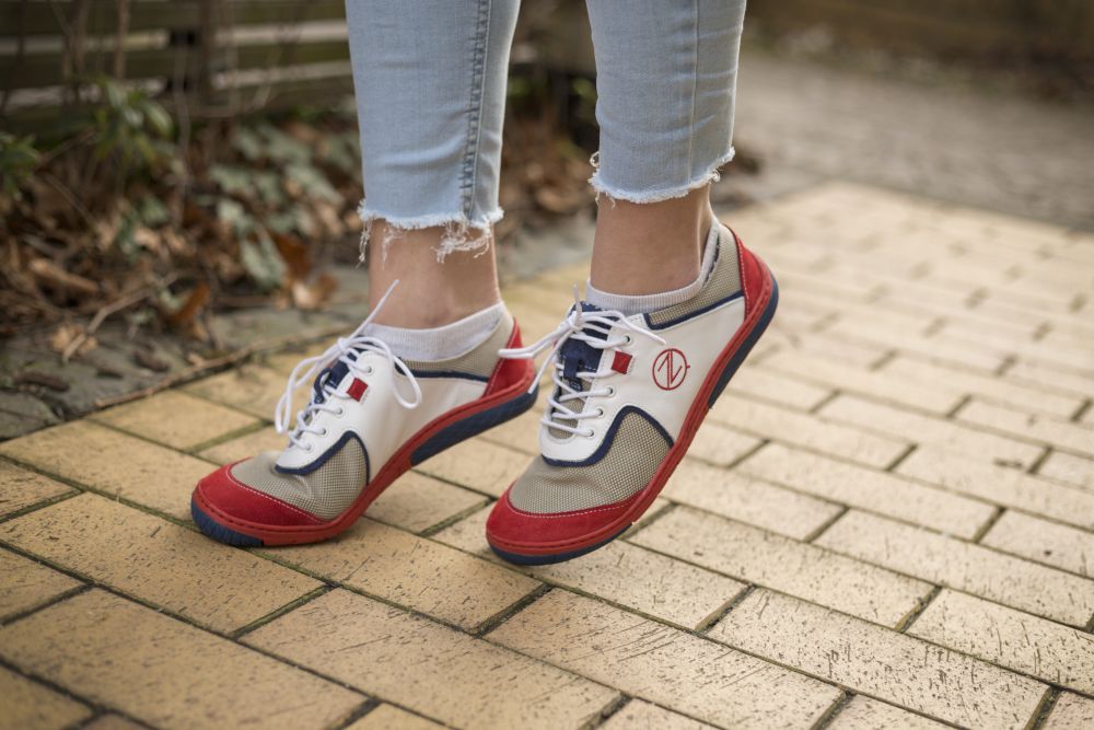 Eine Person trägt die stylischen weißen QOLETTE-Sneaker von ZAQQ und läuft auf einem Ziegelweg, umgeben von üppigen Pflanzen und Grün. Dazu trägt sie hellblaue Jeans mit ausgefranstem Saum.