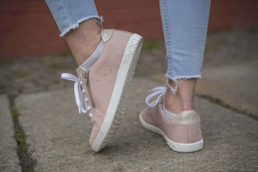 Eine Person in den eleganten QOMFY Wide Rosa Sneakers von ZAQQ läuft einen Steinweg entlang. Die mit einem seitlichen Reißverschluss und glitzerndem Fersendetail verzierten Schuhe zeigen ihr edles Nappaleder-Finish. Hellblaue Jeans mit Fransen an den Knöcheln runden den stylischen Look mit Finesse ab.