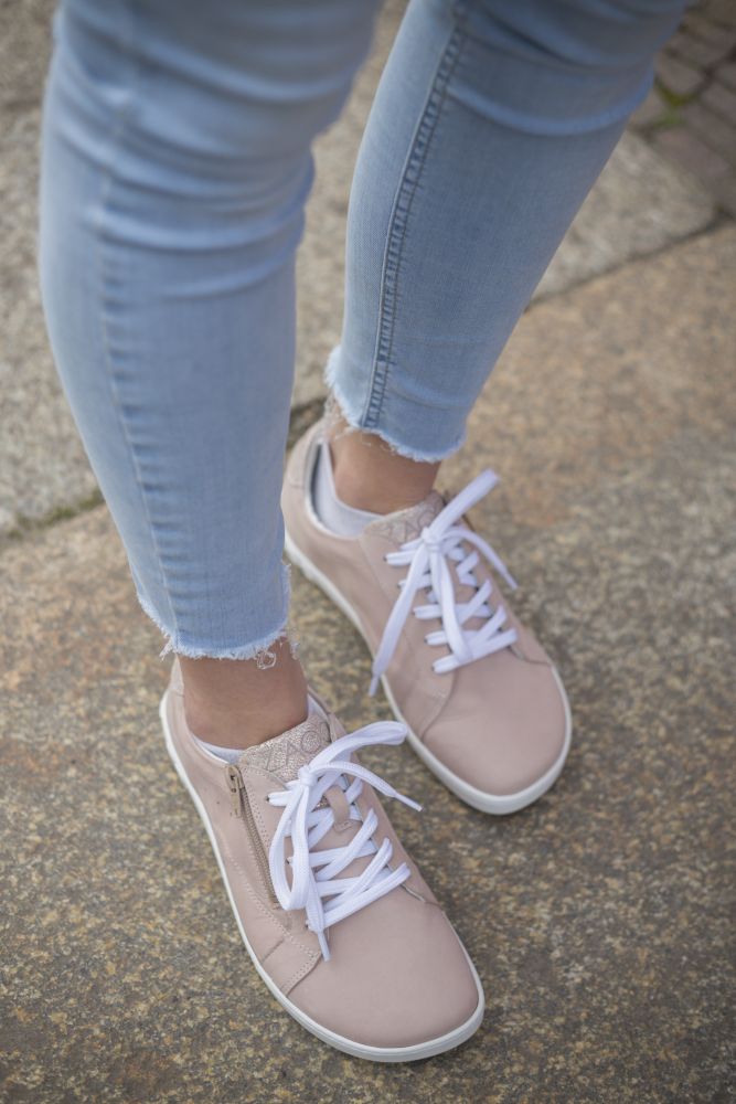 Eine Person in hellblauer Jeans und stylischen ZAQQ QOMFY Wide Rosa Sneakers mit weißen Schnürsenkeln schlendert selbstbewusst über das Steinpflaster.