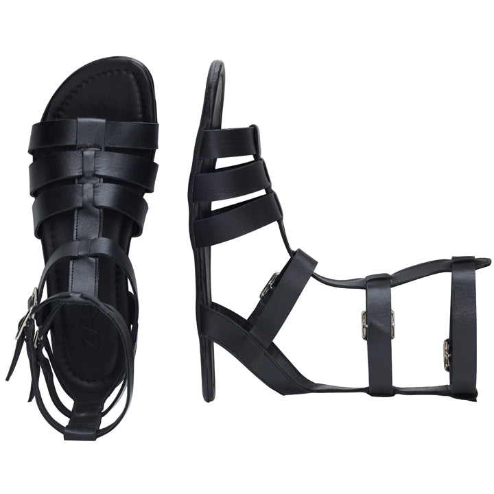 Ein Paar Gladiatorsandalen QORA Black von ZAQQ ist mit mehreren Riemen und Schnallen versehen. Eine Sandale liegt flach, um die Kautschuksohle hervorzuheben, während die andere aufrecht steht und ihre Knöchelhöhe und das detaillierte Riemendesign zur Schau stellt.