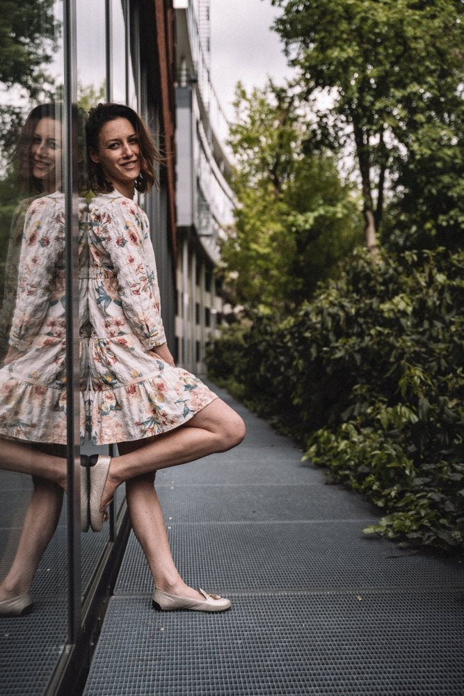 Eine Frau in einem geblümten Kleid und eleganten QOSMO Champagne-Loafern von ZAQQ steht vor einer Glaswand und lächelt, ihr Spiegelbild ist zu sehen. Sie ist draußen, auf einem von Grün umgebenen Weg an einem bewölkten Tag.