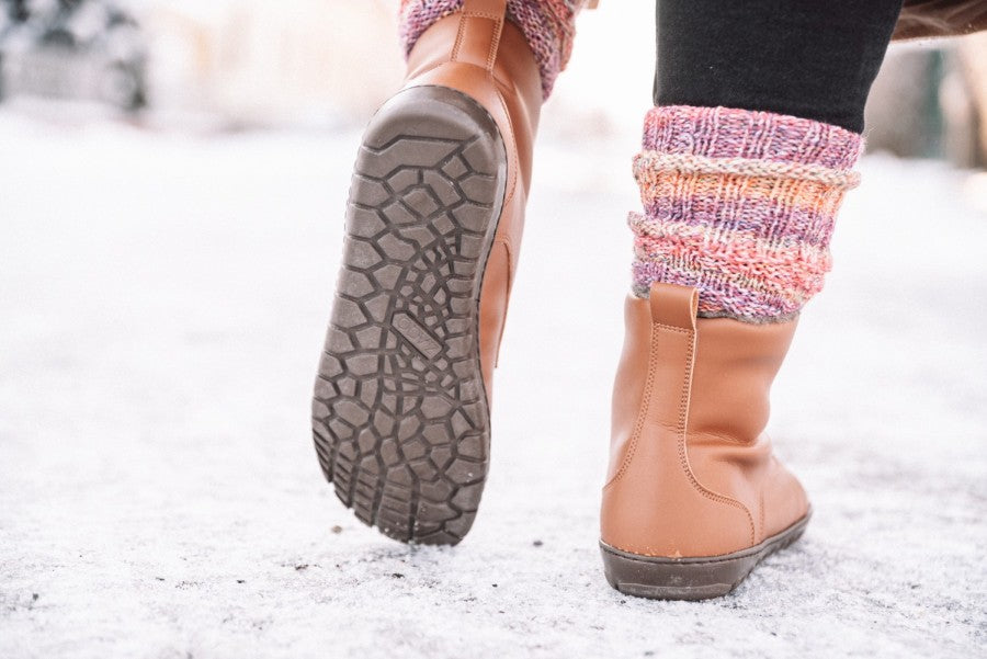 Nahaufnahme einer Person, die auf verschneitem Boden läuft. Präsentiert werden die QUINTIC Winter Vegan Tabaco Stiefeletten von ZAQQ mit ihrer robusten, veganfreundlichen Kautschuksohle und den bunten gestrickten Beinwärmern. Der Hintergrund ist unscharf und betont die kalte, winterliche Umgebung.