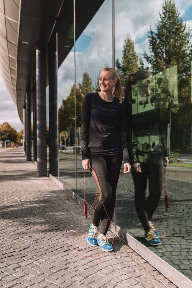 Eine Person in Sportkleidung steht vor einem Gebäude mit reflektierendem Glas und hält ein Springseil. Ihr blau-orangefarbener RIQ-Schuh von ZAQQ liegt auf dem Kopfsteinpflaster. Sie lächelt und schaut zur Seite, im Hintergrund sind ein klarer Himmel und Bäume zu sehen.