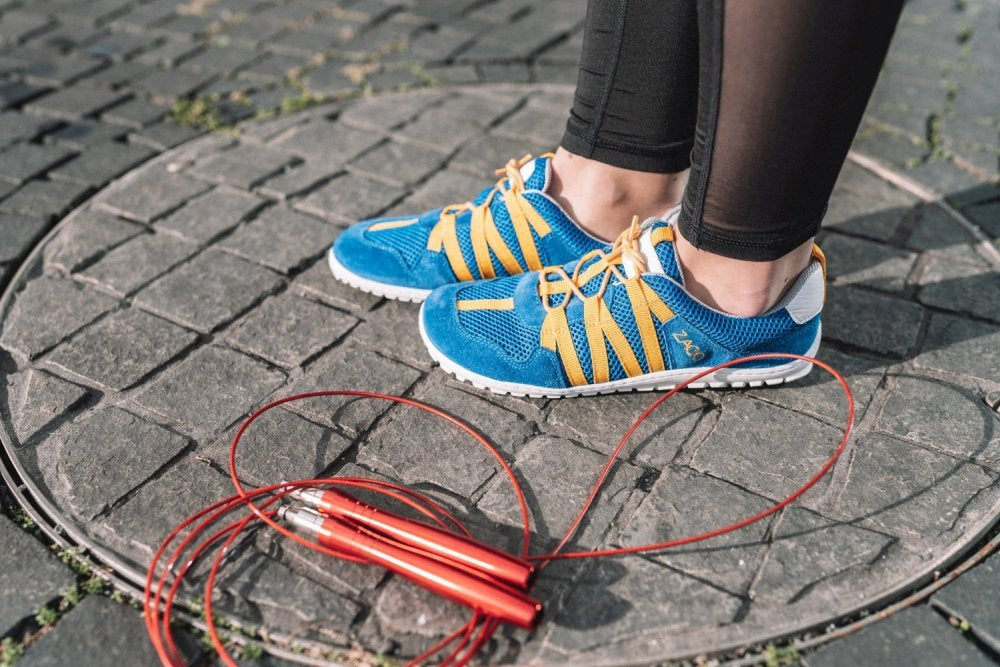 Eine Person trägt ZAQQs Sneaker RIQ Blue Orange mit Veloursleder-Finish und auffälligen gelben Schnürsenkeln und steht auf einem Steinpflaster. Daneben liegt ein aufgerolltes rotes Springseil mit schwarzen Griffen. Schwarze Leggings komplettieren das Ensemble und perfektionieren den sportlichen Look.