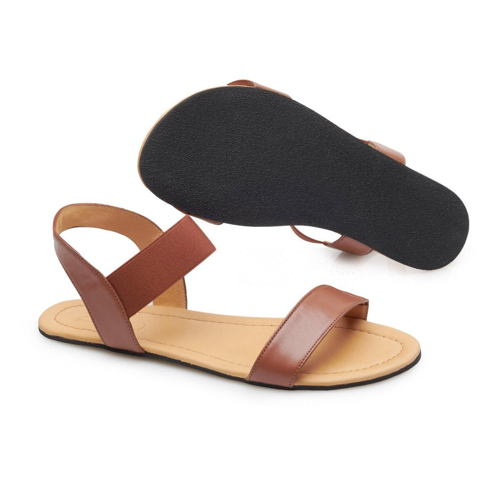 Ein Paar elegante braune ZAQQ SLIQ Cognac-Sandalen mit minimalistischem Design. Eine Sandale ist flach gelegt, um die Innensohle hervorzuheben, während die andere schräg steht, um die schwarze Kautschuksohle zu präsentieren. Diese Sandalen im Barfußstil haben einen Riemen an der Rückseite und einen einzelnen Riemen über den Zehen für zusätzlichen Komfort.